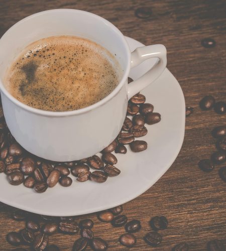 A steaming espresso served in a white cup surrounded by roasted coffee beans on a wooden table.